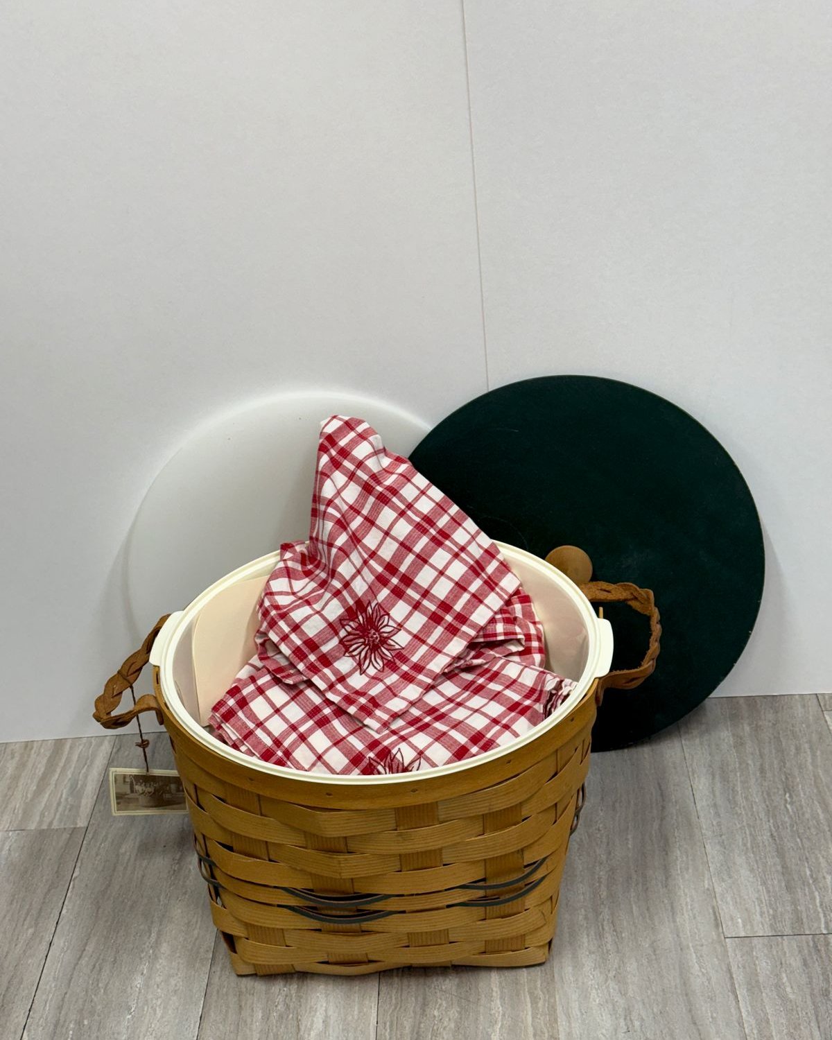 Vintage Peterboro handwoven wood picnic basket with braided leather handles, red and white gingham linens, and a round green lid.