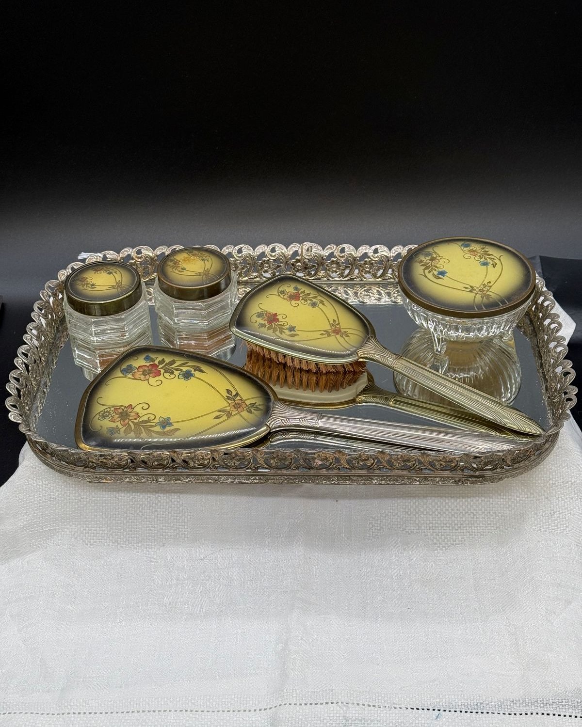 Vintage five-piece vanity set with hand mirror, brushes, and glass jars with floral celluloid backs, arranged on an ornate silver mirrored tray.