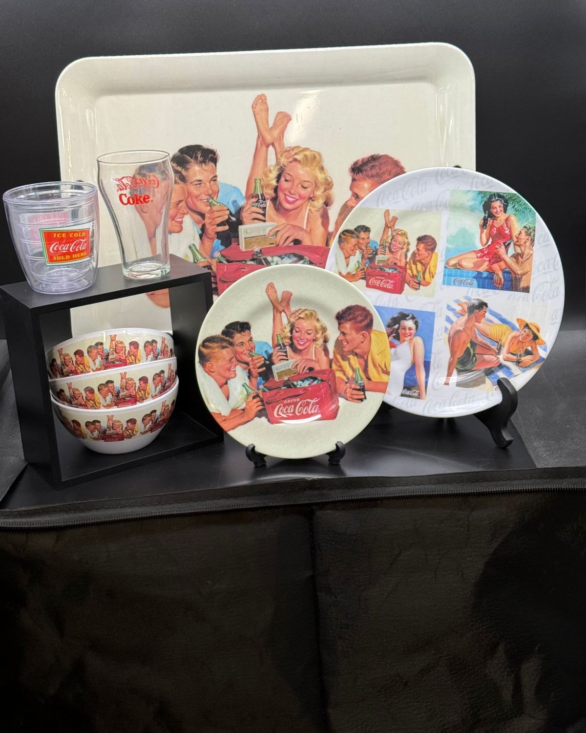 A grouping of Coca-Cola collectibles: a serving tray, two plates, a stack of bowls, a fountain glass, and an insulated tumbler, all with 1950s-style beach and picnic art.
