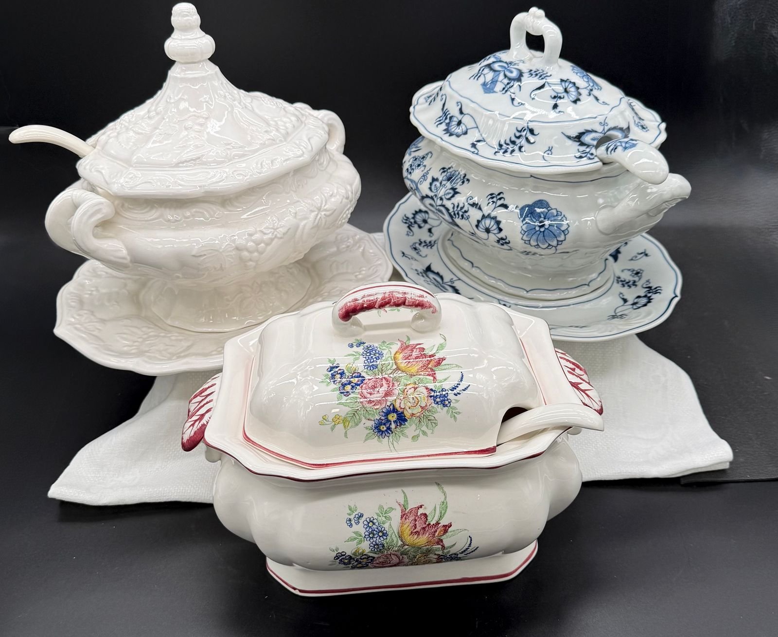 Three covered soup tureens with ladles: a white embossed tureen, a blue and white onion-pattern tureen, and a floral transferware tureen, on underplates.