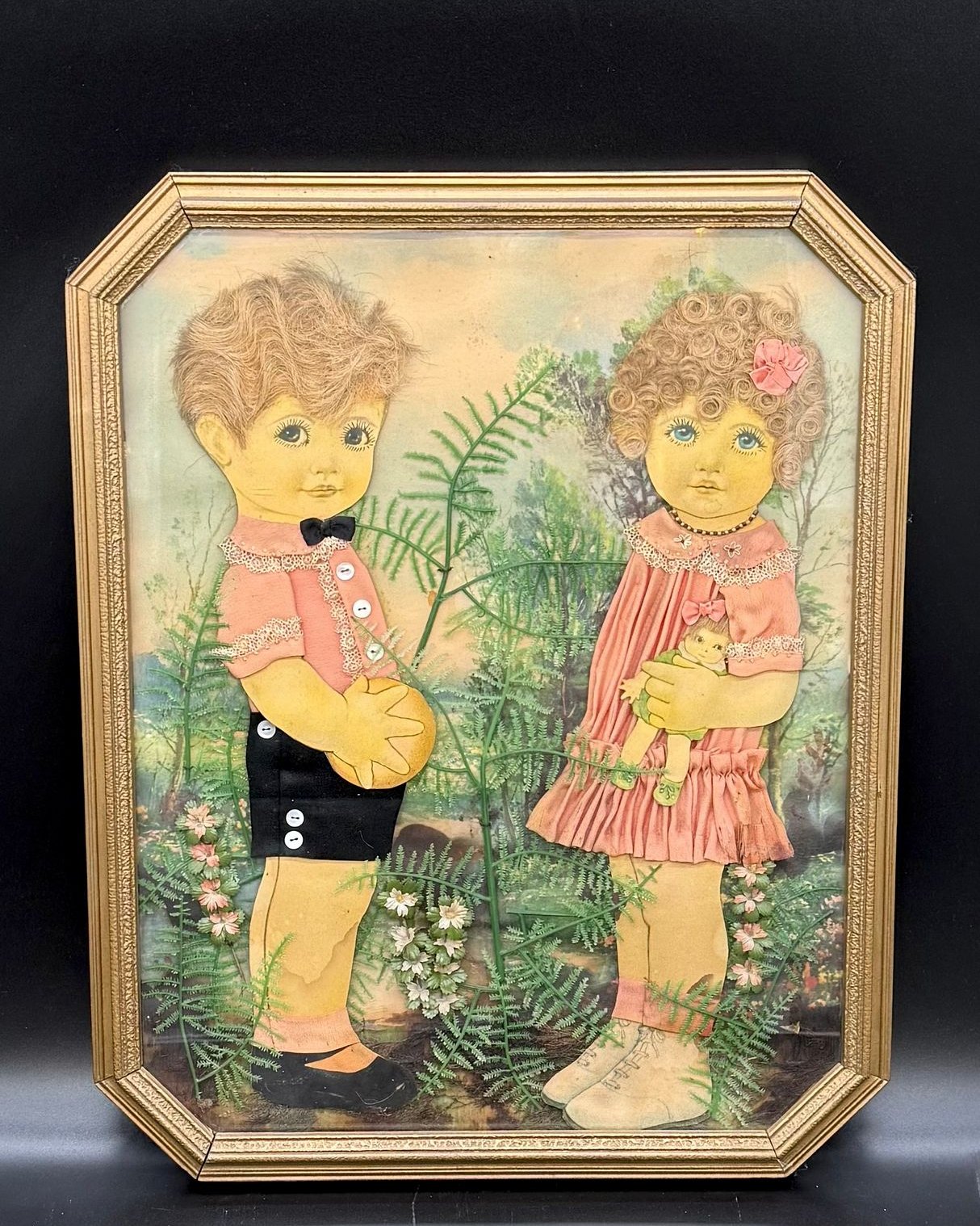 A 16 by 20 inch antique relief picture in an octagonal gilt frame, showing a boy and a girl built from applied fabric with painted faces and real hair, standing in a woodland scene made of real pressed ferns and dried flowers over a painted landscape.