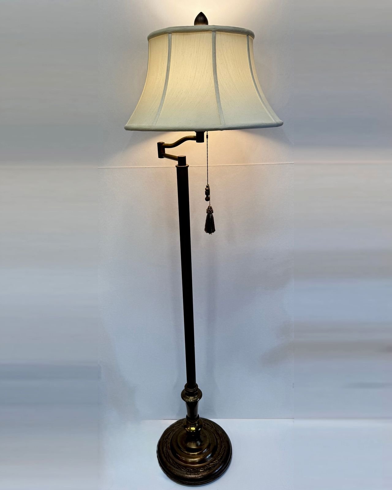 A fifty-six inch vintage brass swing arm floor lamp with a fluted column, a heavy round base cast with a ring of laurel leaves, an acorn finial, a cream bell shaped shade with a scalloped hem, and a pull chain ending in a tassel.