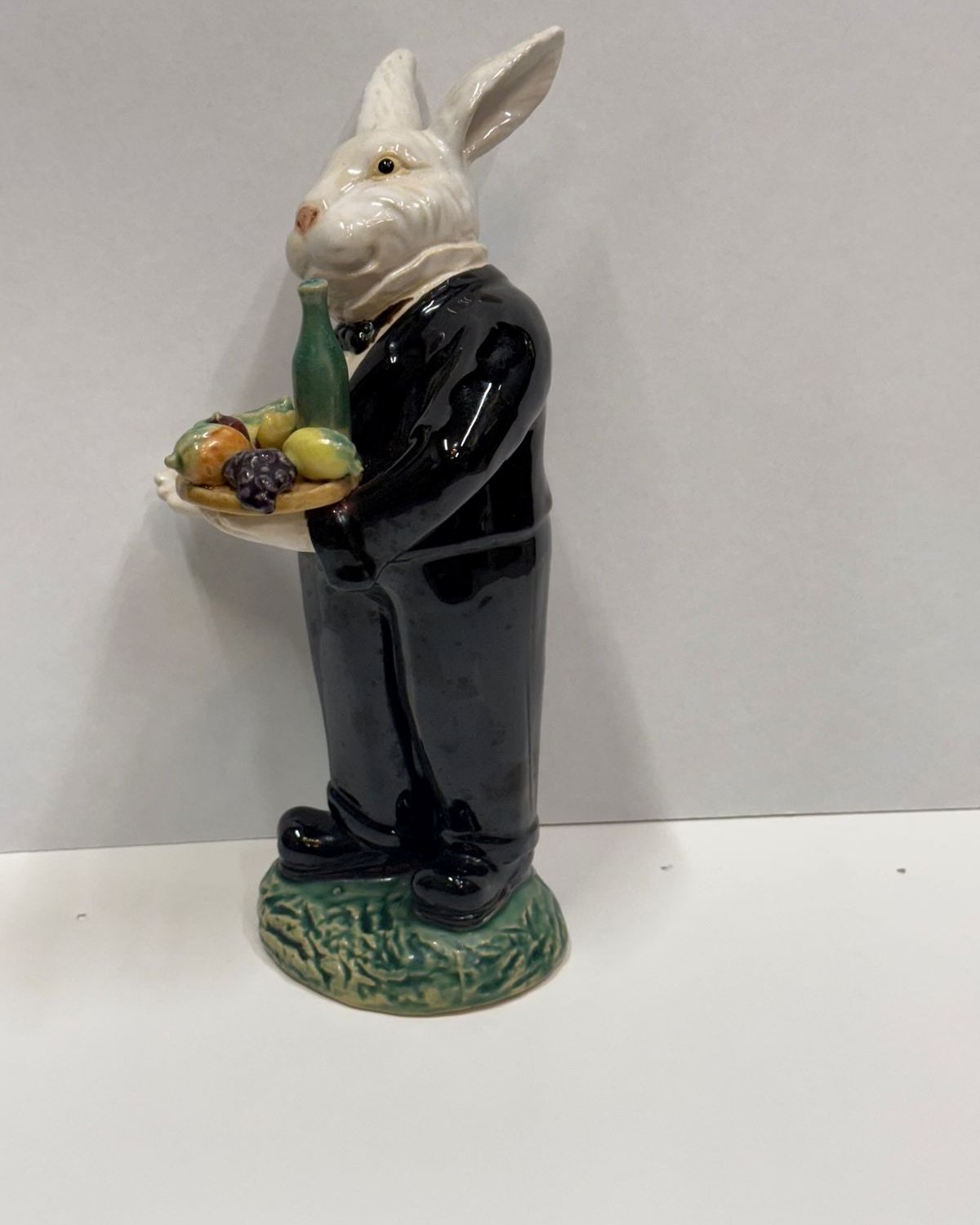 Ceramic figure of a white rabbit in a glossy black tuxedo and bow tie, holding a tray of glazed fruit and a green bottle, standing on a green base.
